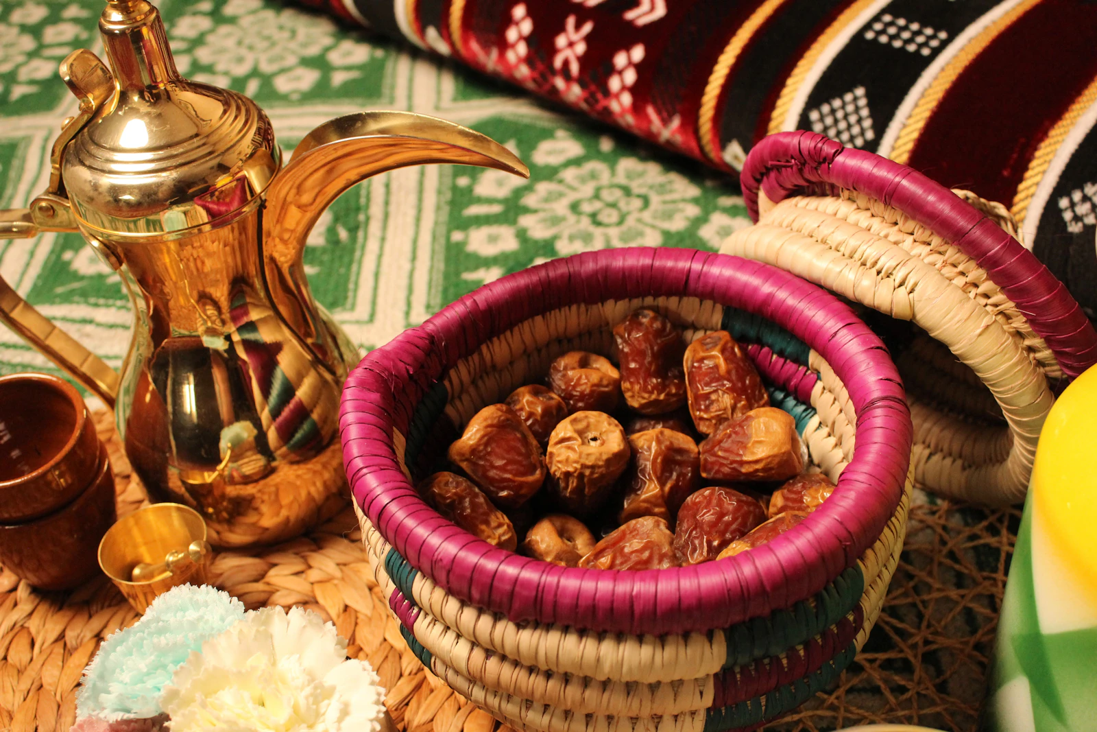 A brass Arabic dallah coffee pot and a woven basket of dates set on a traditional patterned textile.