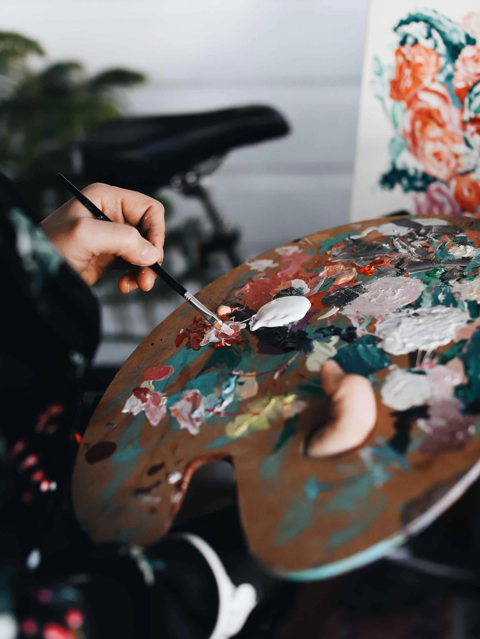 An artist holding a fine brush over a wooden palette loaded with mixed paint, a canvas in progress behind.