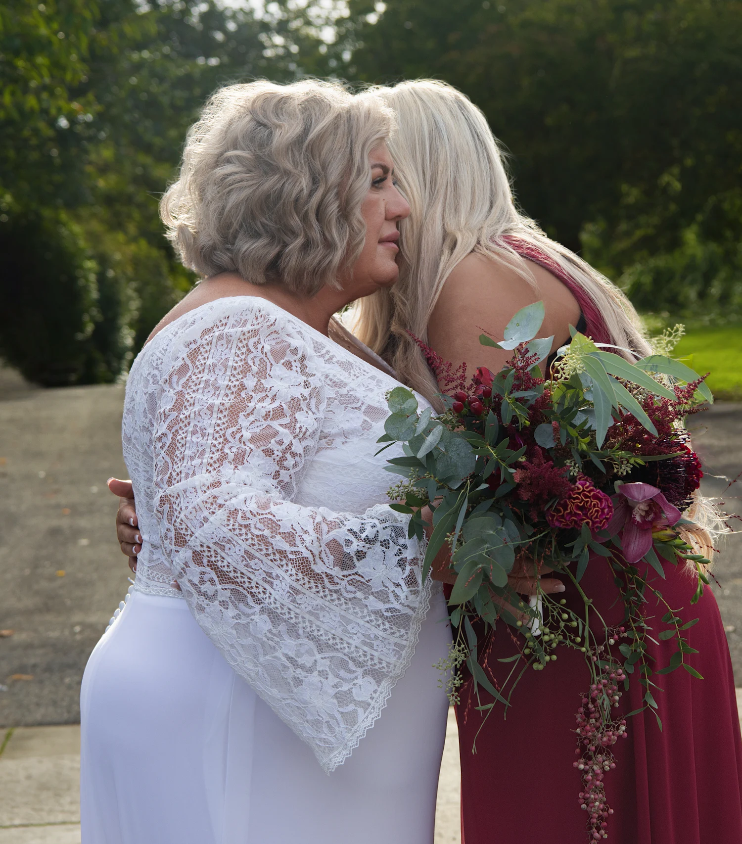 A bride in a white lace gown embracing an older woman holding a bouquet of deep red roses.
