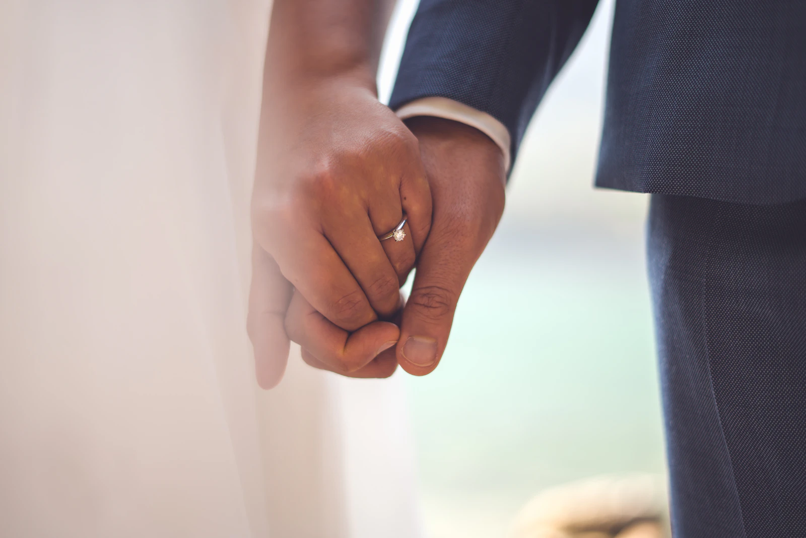 A man in a dark suit and a woman in a white dress holding hands closely, a wedding ring visible on her hand.