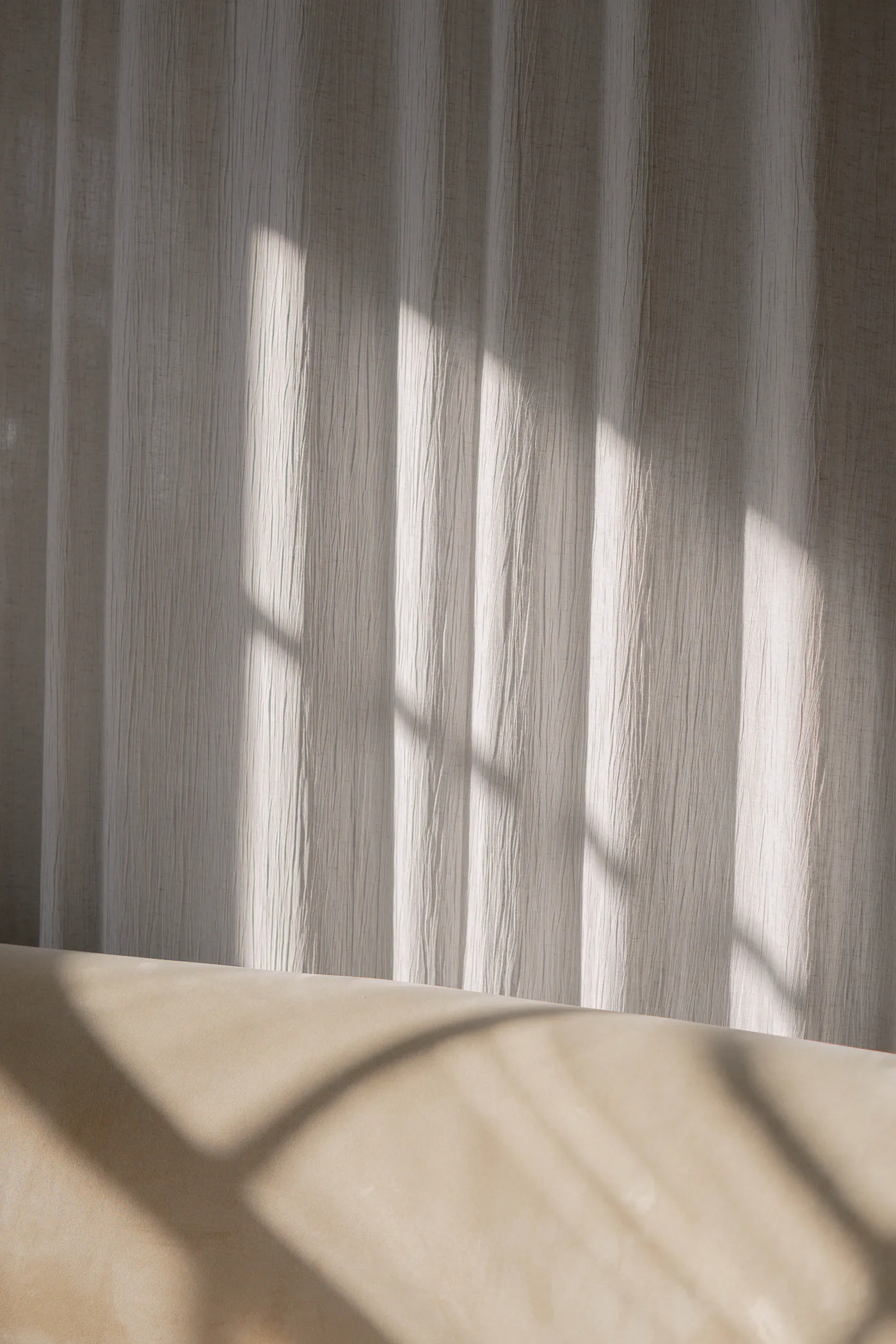 Afternoon sunlight falling through a sheer linen curtain, casting soft window shadows across a pale wall.