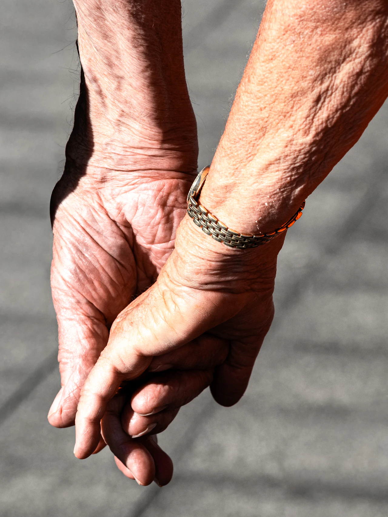 An older couple&rsquo;s clasped hands in warm sunlight, one wrist wearing a metal bracelet.