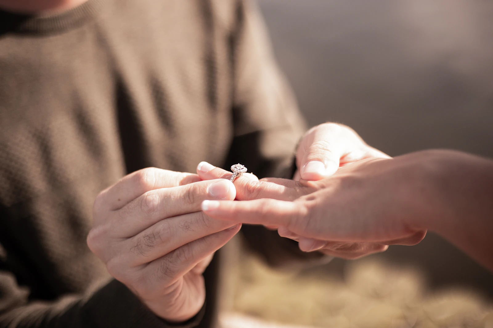 Two hands and a solitaire diamond engagement ring held gently in soft, warm light.