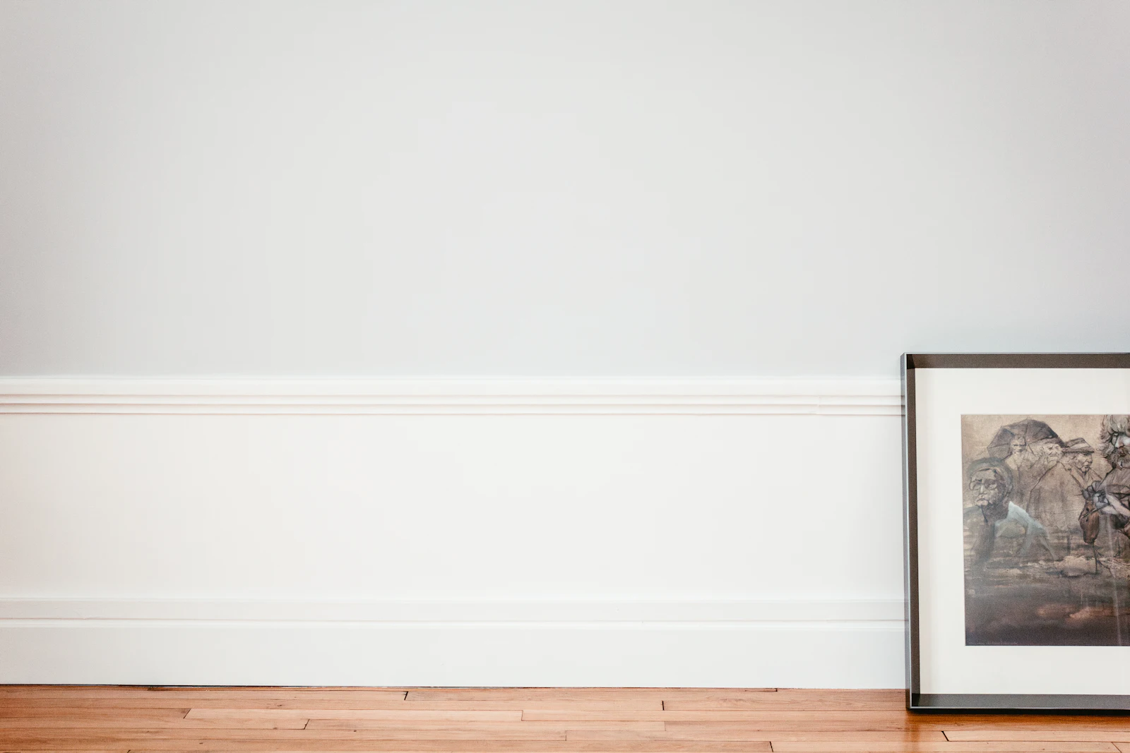 A framed picture leaning against the base of a large empty pale wall above a wooden floor, most of the wall unoccupied.