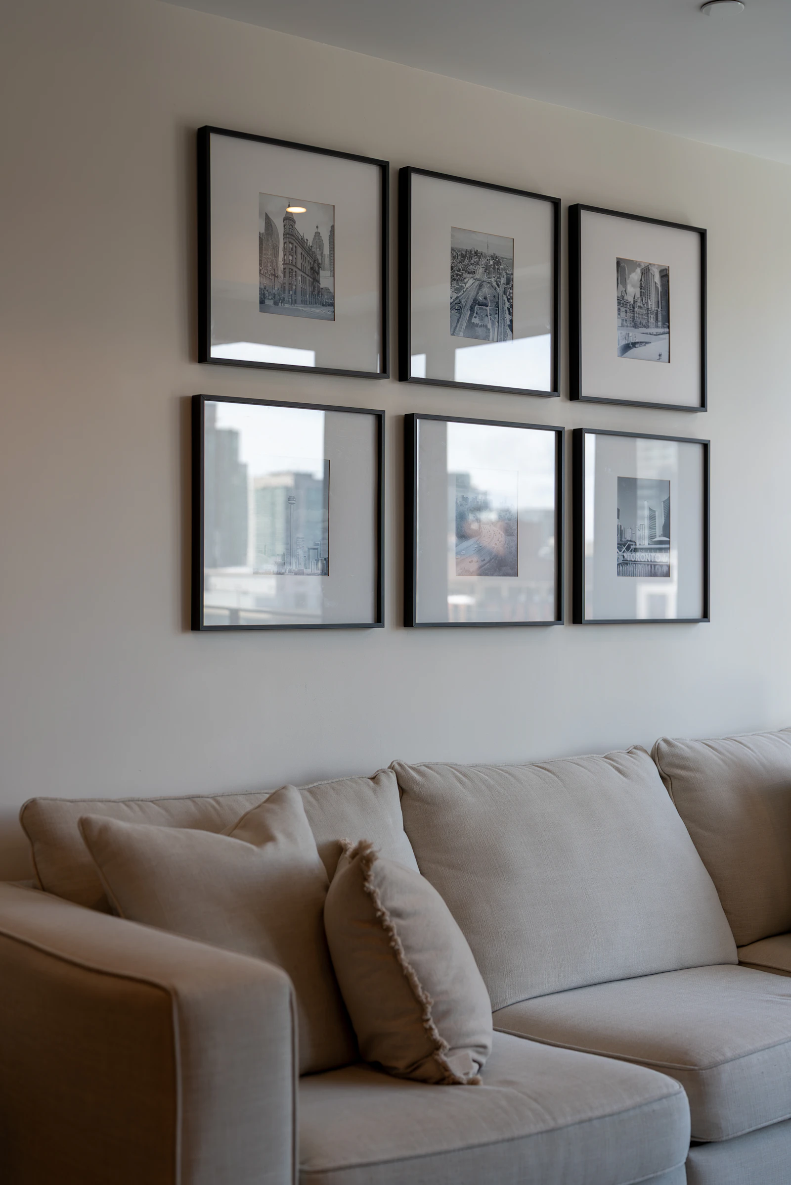 A neat arrangement of framed artworks hung in two rows on a pale wall above a sofa in a warm living room.