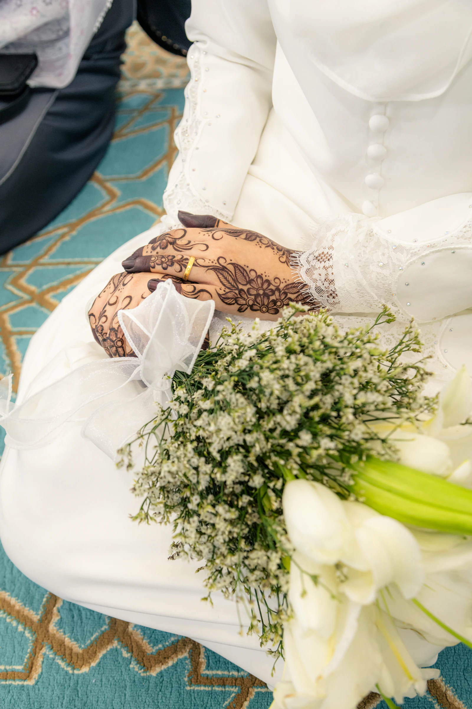 A bride in a white gown with her hands intricately decorated in henna, resting in her lap beside a bouquet of white flowers.