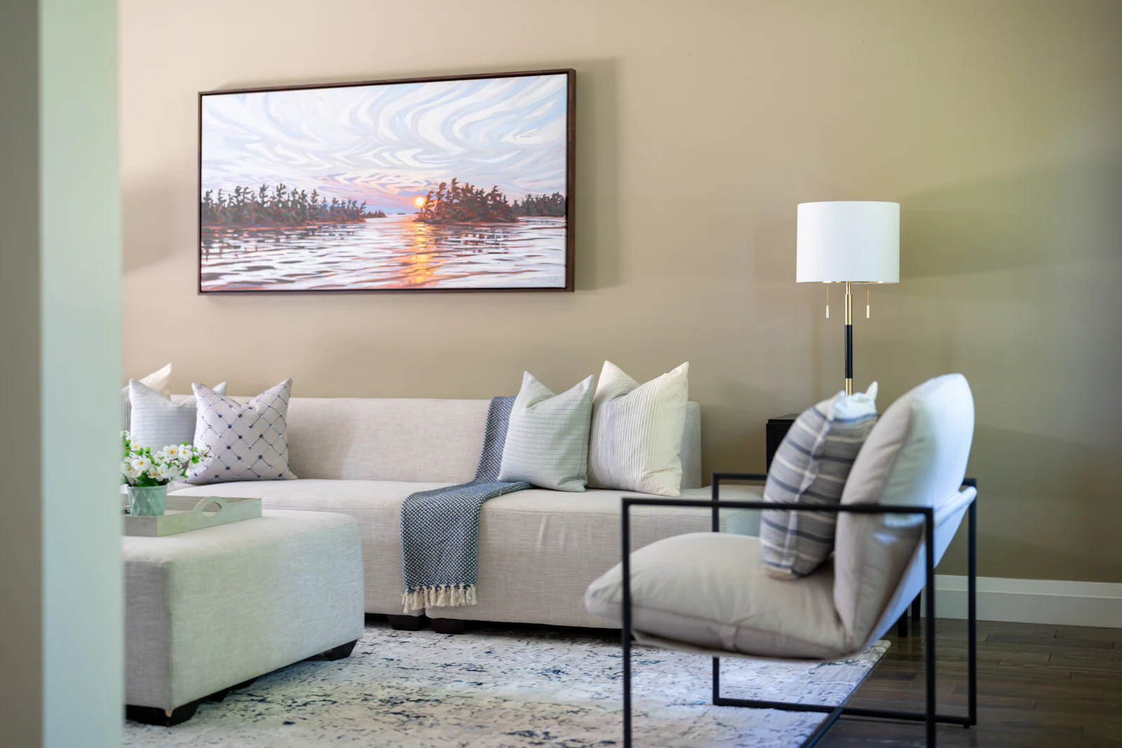 A single large framed landscape painting hung above a cream sectional sofa in a warmly lit living room with beige walls.