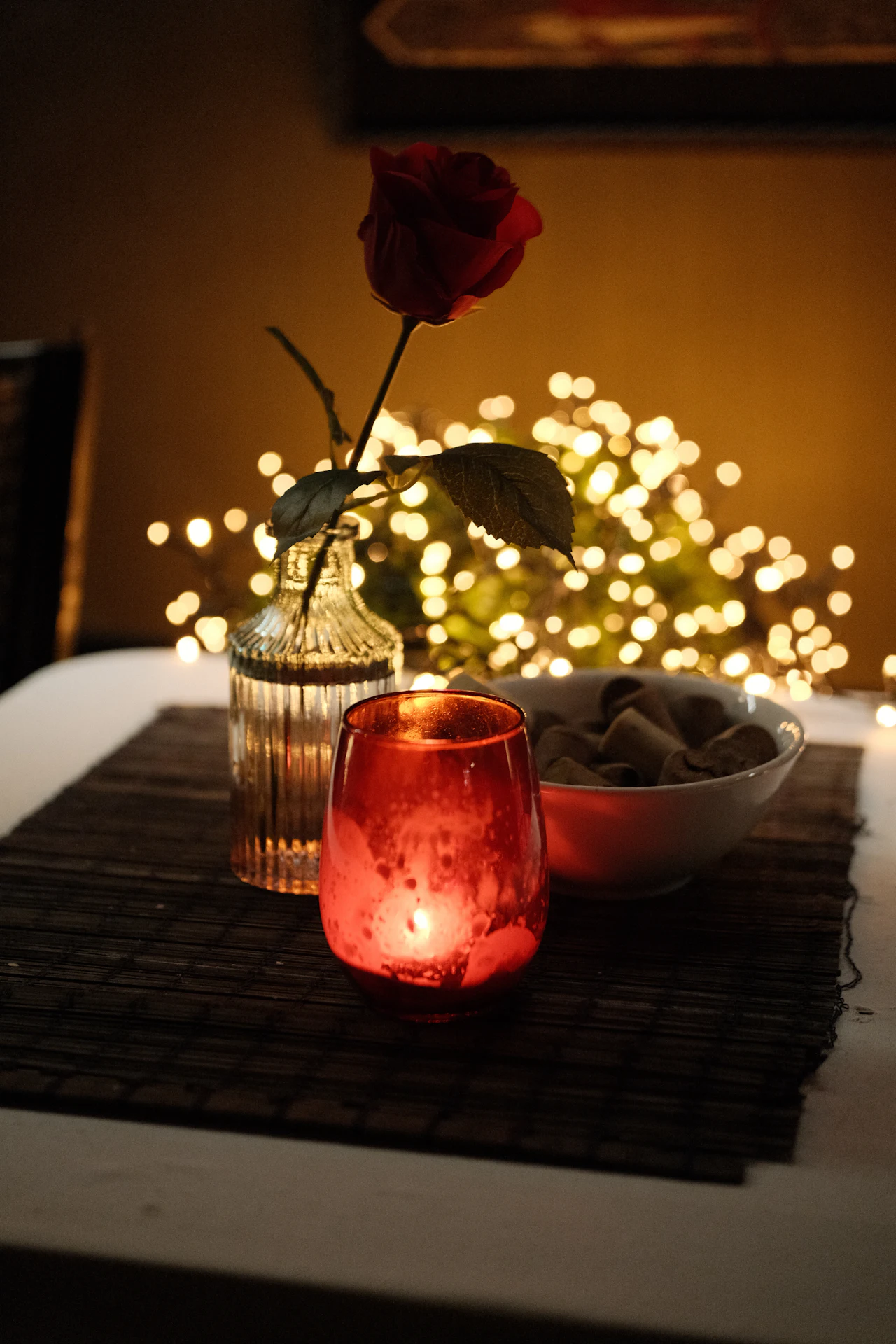 A single red rose in a glass vase beside a lit red votive candle, with warm fairy-light bokeh behind.